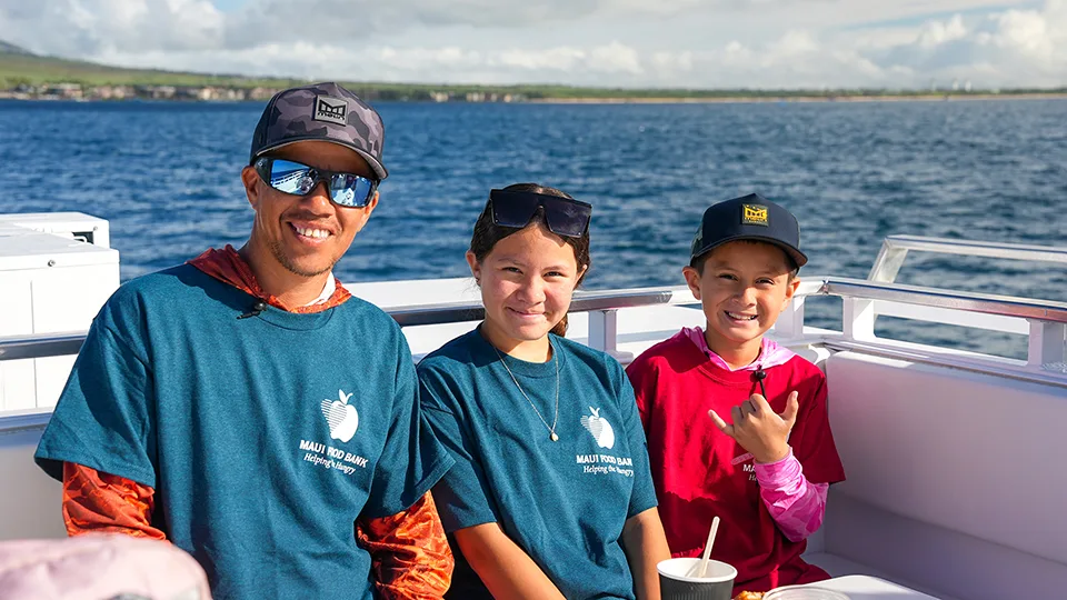 Maui Food Bank team members in blue t-shirts with child in red shirt smiling on Pride of Maui recognition cruise
