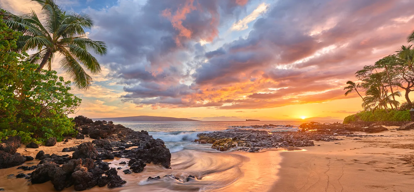 Sunset at Secret Beach in South Maui with lava rocks, golden sand, palm trees, and vivid orange clouds reflecting over the ocean.