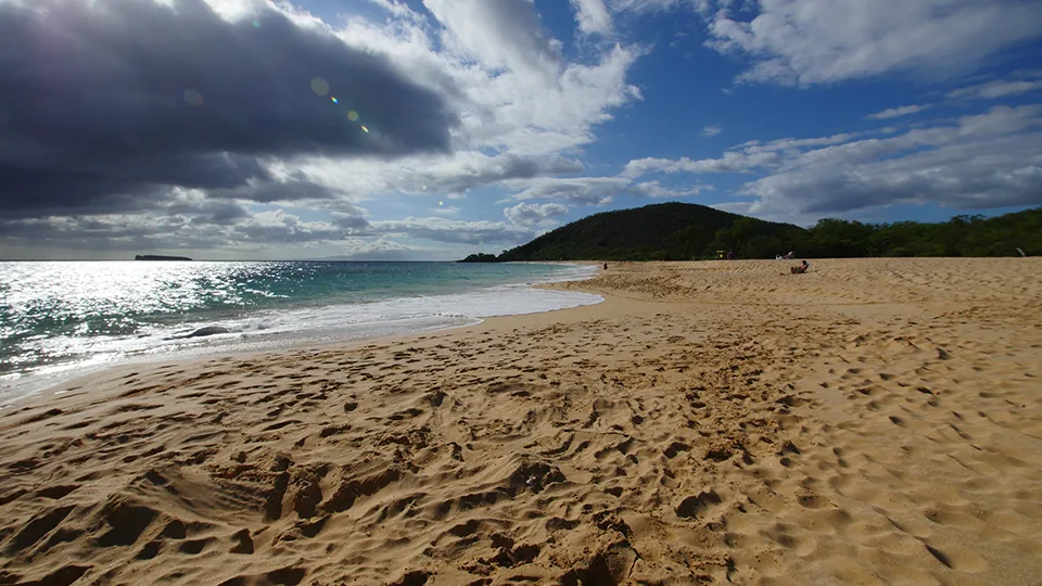 View of Big Beach shoreline with soft waves and rocky headland, captured under dramatic clouds.