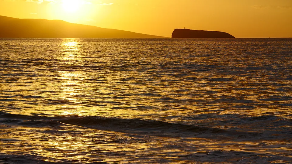 Molokini Crater silhouetted at golden hour from the shore of Poʻolenalena Beach.
